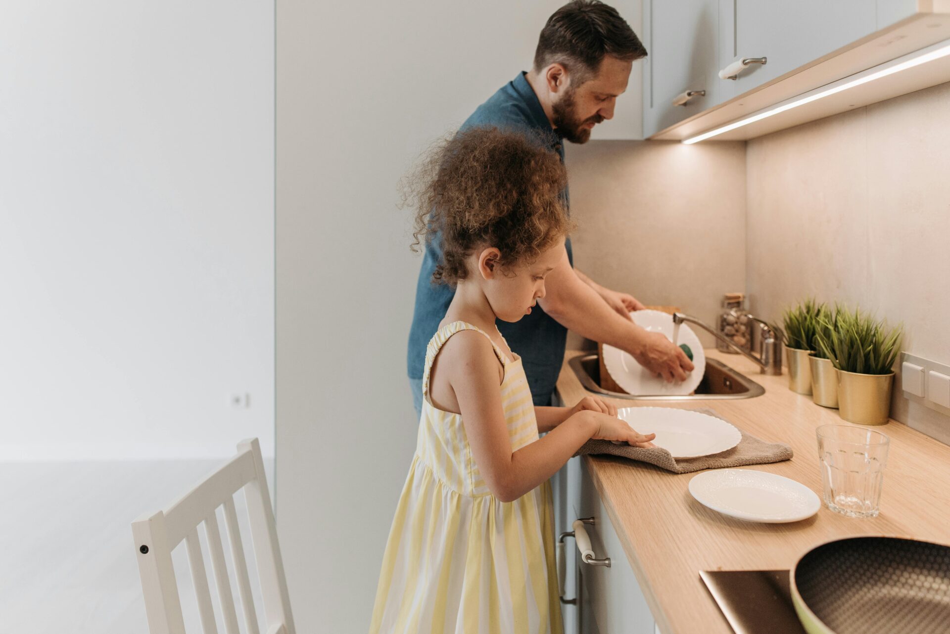 A father and daughter wash dishes in the kitchen with functionality given by under-cabinet lights.